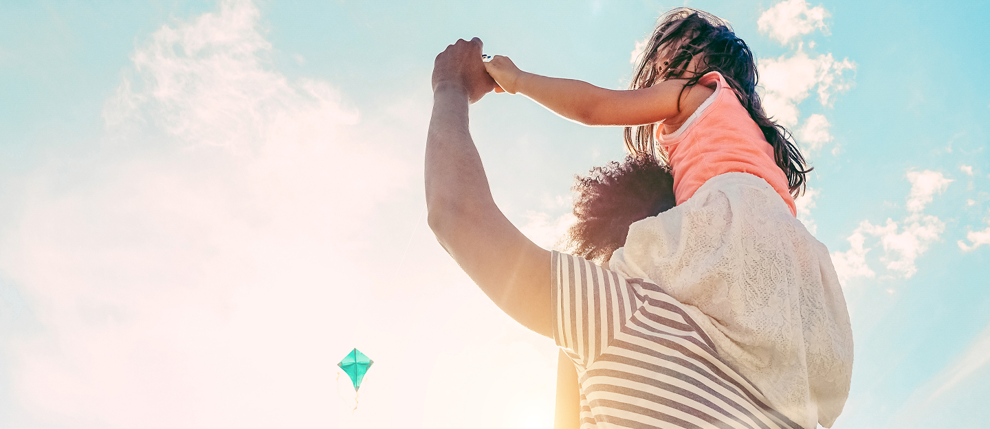 Man with daughter sitting on his shoulders stares at sky with kite in the background